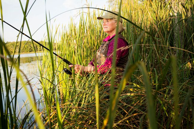 A woman casts a fishing rod along a reedy shorelin